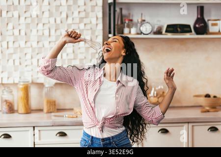 Donna in cucina che canta mentre tiene in mano una frusta e balla felicemente durante il tempo di cottura Foto Stock