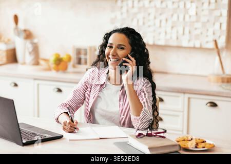Donna che parla al telefono mentre si siede a una scrivania con un computer portatile e spuntini in cucina Foto Stock