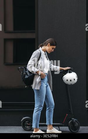 Foto a tutta lunghezza di giovane donna d'affari in piedi in un edificio con scooter elettrico a spinta verso il basso Foto Stock