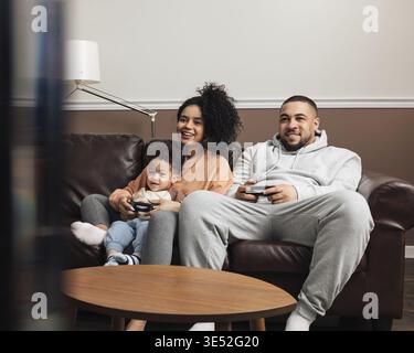 Madre, padre e figlio piccolo che giocano a videogiochi mentre si siedono sul divano a casa Foto Stock