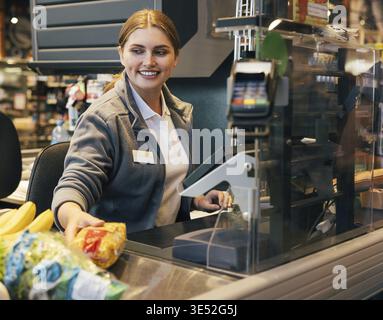 Cassiera sorridente che lavora al registratore di cassa e sposta i prodotti scansionati Foto Stock