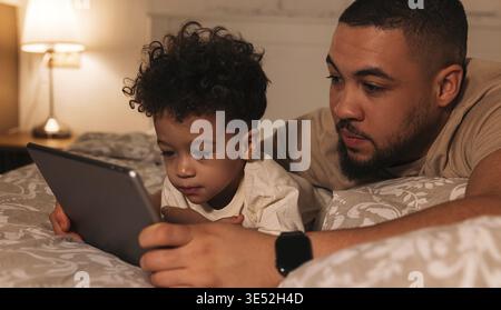 Padre e figlio guardano il tablet digitale mentre si sdraiano a letto la sera Foto Stock