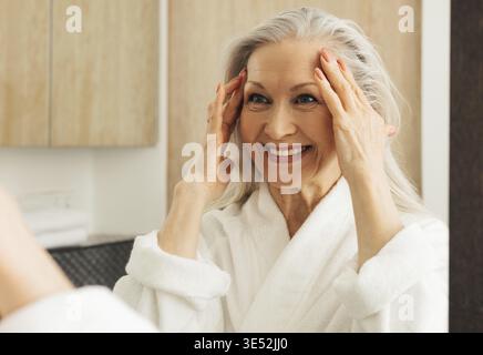 Donna anziana felice con i capelli grigi che guardano uno specchio e che toccano il suo volto mentre guarda lo specchio Foto Stock