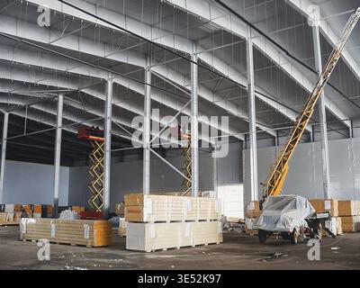 Cantiere con elevatori a forbice, materiali da costruzione e un camion con gru all'interno di un grande magazzino Foto Stock
