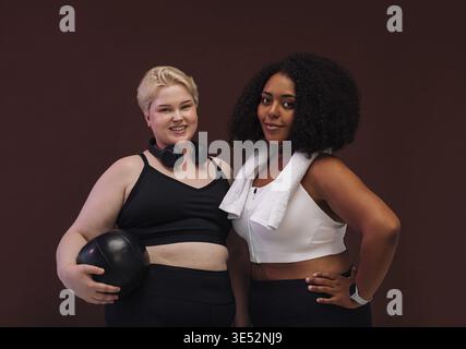 Due allegre donne in abbigliamento sportivo in posa su sfondo marrone. Inoltre, le donne di taglia superiore sono in piedi insieme alle attrezzature per il fitness Foto Stock