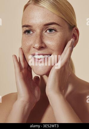 Ritratto molto dettagliato di una giovane donna sorridente che si massaggia il viso. Primo piano di una donna bionda che tocca la sua pelle lentigginita Foto Stock
