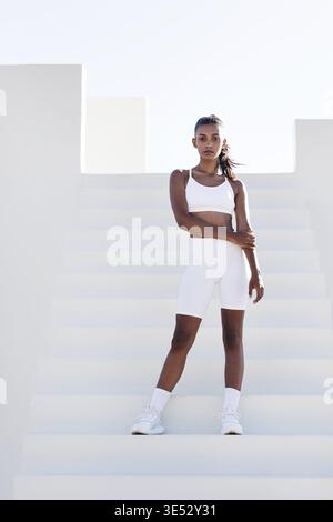 Donna slim-fit a tutta lunghezza con abbigliamento sportivo bianco. Donna in piedi su scale bianche che si rilassa durante l'allenamento Foto Stock