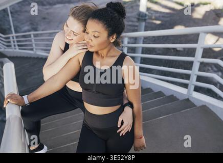 Due amiche in un abbigliamento da fitness nero in piedi insieme sulle scale. Due diversi atleti si rilassano dopo l'esercizio fisico Foto Stock