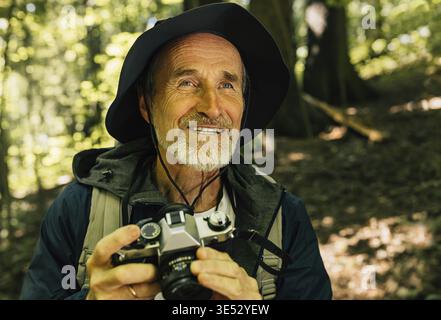 Ritratto di un maschio sorridente con una macchina fotografica. Uomo anziano seduto nella foresta e a scattare foto con la sua macchina fotografica Foto Stock