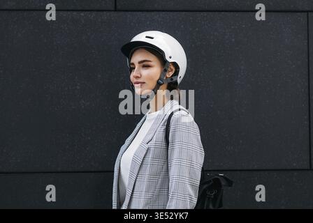 Donna d'affari in casco bianco da ciclismo con zaino in piedi contro una parete nera all'aperto Foto Stock