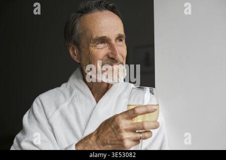 Primo piano di un uomo maturo sorridente che indossa un accappatoio bianco che tiene in mano wineglass Foto Stock