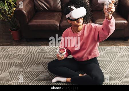 Donna sorridente in salotto con VR set. Giovane donna in abiti comodi che gioca a videogiochi mentre si siede Foto Stock