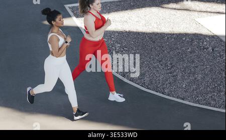 Ripresa dall'alto di due donne che corrono insieme all'aperto. Angolo elevato di due amiche che fanno jogging insieme indossando abbigliamento sportivo in diversi colori Foto Stock