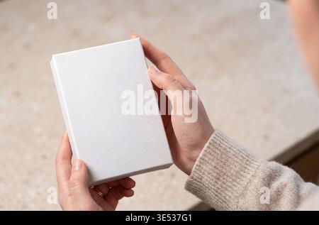 Mockup delle mani di una persona che tiene una piccola scatola bianca vuota su una superficie di pietra testurizzata. Foto Stock