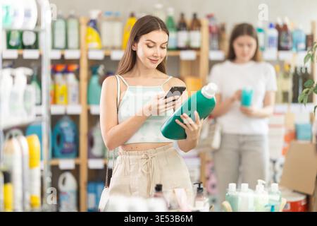 La donna esegue la scansione del codice QR sull'etichetta del detergente utilizzando il suo smartphone, mentre gli acquirenti in background sfogliano i prodotti Foto Stock