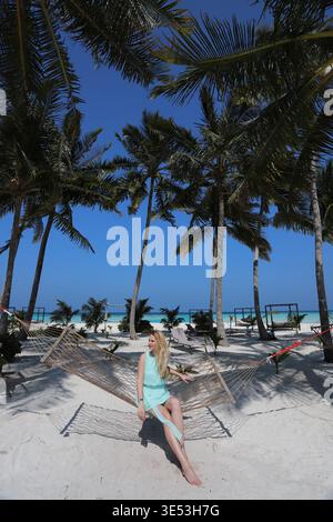Donna bionda sorridente in abito turchese che si rilassa sull'amaca di corda sotto le alte palme di cocco sulla spiaggia di sabbia bianca, lussuoso resort delle Maldive Foto Stock