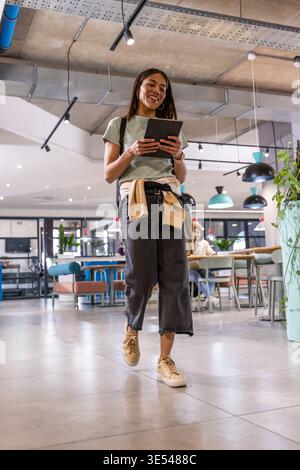 Donna che cammina attraverso lo spazio di lavoro condiviso tenendo un tablet, sorridente, indossando una T-shirt verde e uno zaino Foto Stock