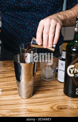 Di persona che versa liquido scuro dal jigger allo shaker da cocktail su un tavolo di legno accanto alla bottiglia Cynar e agli amari Angostura. Cynar, Angostura, mixologia Foto Stock