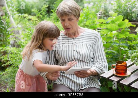 nonna e piccola nonna che usano il tablet e si divertono insieme in giardino. Famiglia felice che ama l'estate all'aria aperta. || modello rilasciato Foto Stock