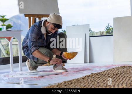 Uomo barbuto accovacciato su un foglio in studio, indossando un berretto, applicando vernice dalla ciotola alla tela del pavimento. Artista, minimalista, luce del giorno, finestra, cavalletto, tappeto, cuffie || modello rilasciato Foto Stock
