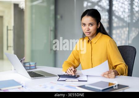 Una giovane ragioniera che indossa una camicia gialla e calcola i dati finanziari utilizzando una calcolatrice e documenti, mentre lavora alla sua scrivania in un ambiente d'ufficio moderno Foto Stock