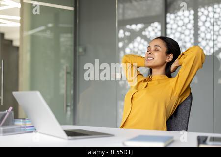 Contenta una giovane donna d'affari che gode di un momento di relax e pace, si allunga alla scrivania dell'ufficio con un laptop, trova equilibrio e sollievo dallo stress durante una giornata di lavoro intensa Foto Stock