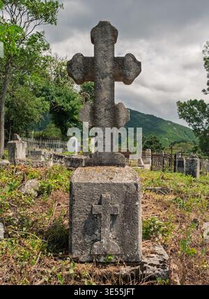 L'antico monumento a croce in pietra sorge sopra l'erba ricoperta in un tranquillo cimitero rurale in un pomeriggio nuvoloso Foto Stock