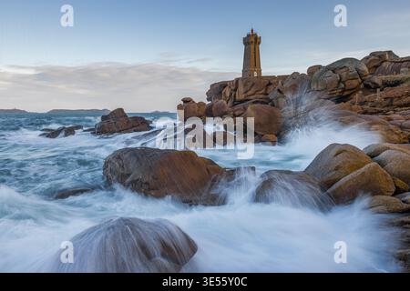 Veduta del faro di Ploumanac'h in piedi su rocce di granito rosa con onde che si infrangono sotto un morbido cielo blu a Perros-Guirec, Bretagna, Francia. Foto Stock