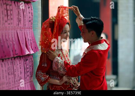 Giovane coppia vietnamita vestita con costumi da sposa in stile cinese rosso posa vicino a tavolette di preghiera. Ho chi Minh. Vietnam. Foto Stock