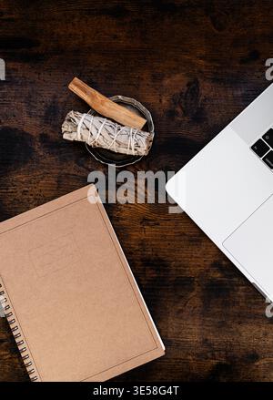 Notebook, laptop e palo santo con salvia bianca su tavolo in legno, spazio di lavoro minimo piatto con elementi naturali. Vista dall'alto. Copia spazio Foto Stock