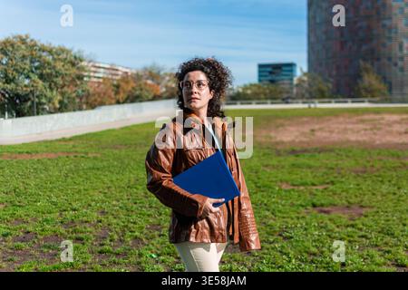 Donna d'affari con capelli ricci che indossa una giacca di pelle marrone e occhiali, che cammina in un parco urbano verde tenendo in mano una cartellina blu, con la torre gl Foto Stock