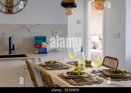 Elegante cucina open space con tavolo da pranzo, piatti freschi, bicchieri da vino e arredamento moderno che creano uno spazio interno caldo e invitante Foto Stock