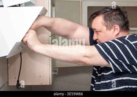 Uomo in camicia a righe lavora su un armadio da cucina, raggiungendo l'interno per regolare una moderna installazione di cappa gamma, con uno sfondo da cucina di colore neutro Foto Stock