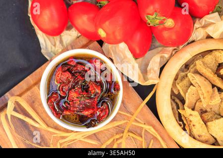 I pomodori freschi riposano accanto a un recipiente di olio di pomodoro essiccato al sole. Pezzi di pane croccanti ti attendono in un recipiente di legno. La luce del sole mette in risalto i colori ricchi su un boa di legno Foto Stock