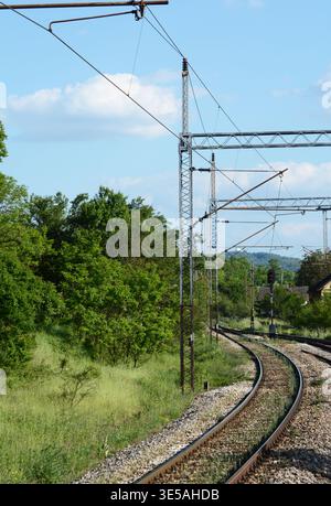 curva ferroviaria, pali metallici con fili elettrici, alberi verdi e cielo blu sopra di essi Foto Stock