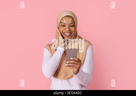 Giovane donna musulmana nera in hijab che reagisce con sorpresa mentre legge lo smartphone Foto Stock