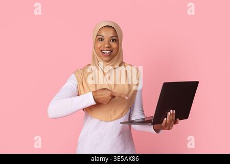 Giovane donna musulmana nera in hijab che punta eccitatamente al portatile in mano Foto Stock