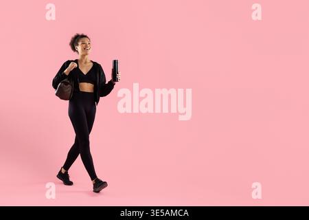 Giovane donna nera sportiva in abbigliamento sportivo nero che cammina con sicurezza, tenendo in mano una bottiglia d'acqua Foto Stock