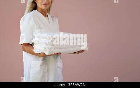 Una donna adulta in uniforme professionale bianca contiene una pila ordinata di asciugamani e biancheria da letto bianchi piegati. Sfondo rosa polveroso uniforme, luce da studio pulita. Foto Stock