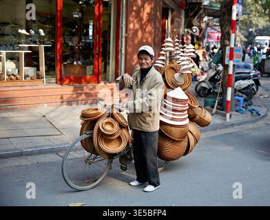 Un uomo sta accanto a una bicicletta carica di vari cesti ad Hanoi. Sorride e ondeggia le persone che passano nelle strade trafficate. Foto Stock