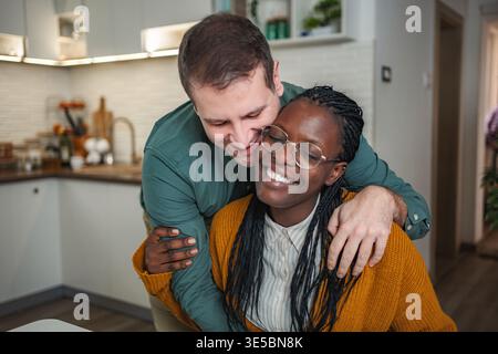 Felice coppia interrazziale che si abbraccia nella cucina di casa Foto Stock