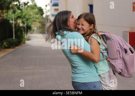Madre che tiene in braccio la figlia con lo zaino in una strada urbana, celebrando il momento del ritorno a scuola Foto Stock