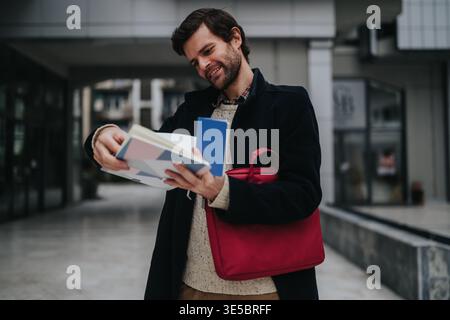 Un giovane uomo d'affari sorridente che legge documenti all'aperto con la borsa rossa per laptop nella piazza urbana degli uffici Foto Stock