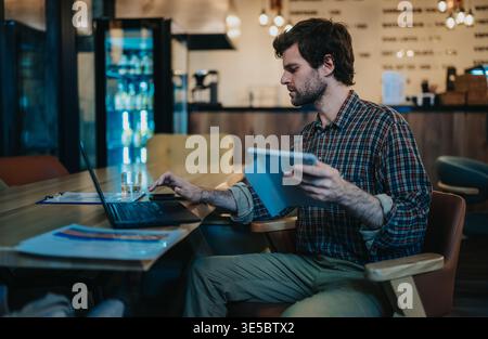 Uomo d'affari che lavora con un notebook e un tablet in un accogliente bar mentre esamina i documenti Foto Stock