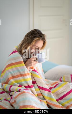 Donna rilassata che beve un caffè seduto su un comodo letto in una camera accogliente con raggi di sole mattutino. Foto Stock