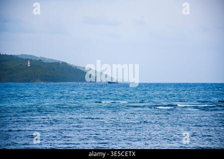 Tranquillo mare blu con onde dolci, una barca da pesca distante e un faro a strisce su un promontorio lussureggiante Foto Stock