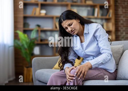 Donna seduta sul divano che stringe il ginocchio nel dolore, strisciando per disagio articolare e infiammazione, raffigurante lesioni croniche o acute alle gambe, artrite o disagio medico a casa Foto Stock