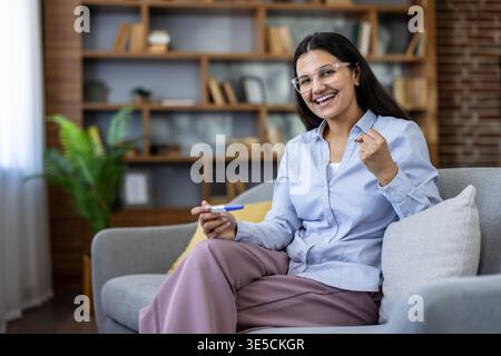 Donna felice seduta su un divano, che tiene un test di gravidanza con un risultato positivo, sorridente e gestante con una pompa a pugno, celebrando buone notizie a casa Foto Stock