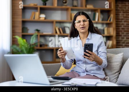 Una giovane donna stressata che sembra confusa mentre si siede su un divano a casa, tiene in mano una ricevuta e uno smartphone, calcola le spese e si trova di fronte a difficoltà finanziarie Foto Stock