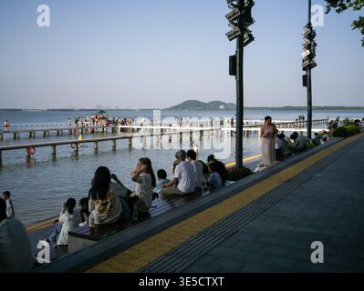 Wuhan, Cina - 16 giugno 2025: Persone sedute su gradini di legno accanto a una passeggiata lastricata sul lungomare con pavimentazione tattile vicino a un lago. L'immagine mostra un lon Foto Stock